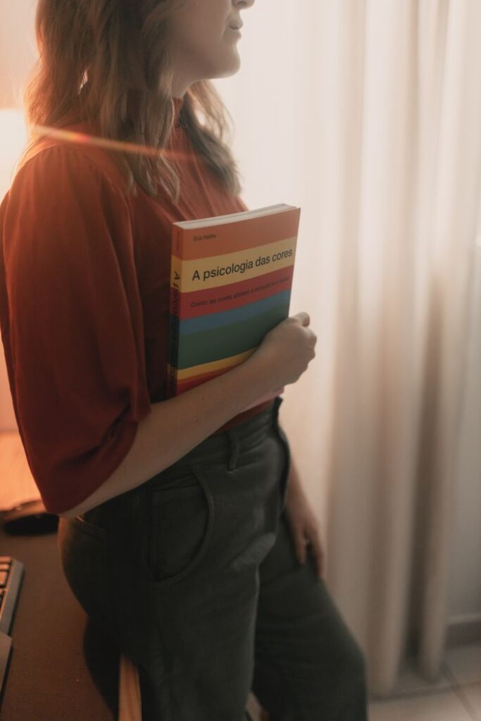 Woman standing indoors holding a book on color psychology, bathed in warm light.